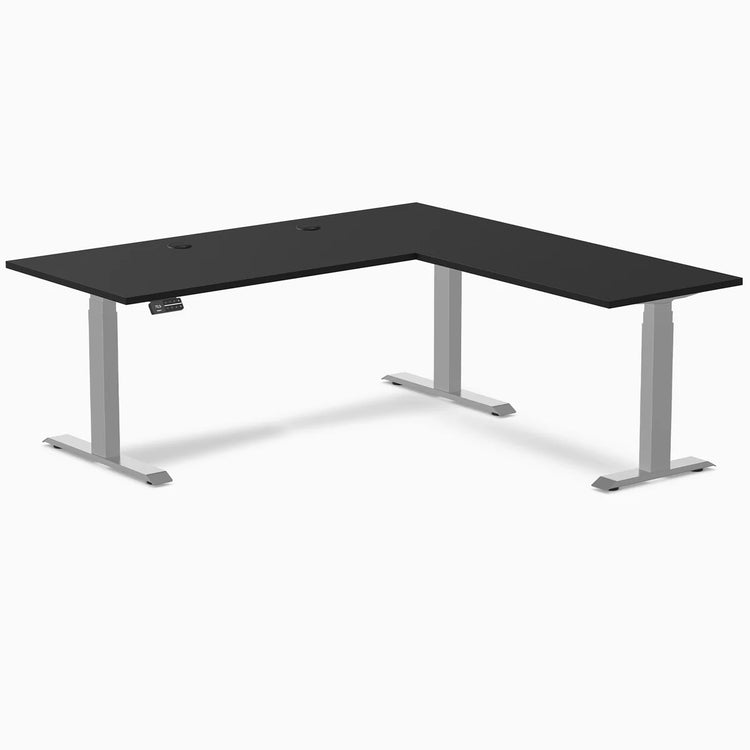 Black laminate L-shape sit-stand desk with gray adjustable legs and grommets.