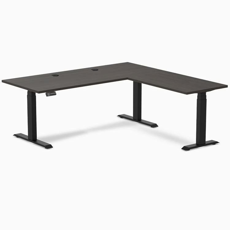Burnished Wood L-shape laminate sit-stand desk with matte black legs and grommets.