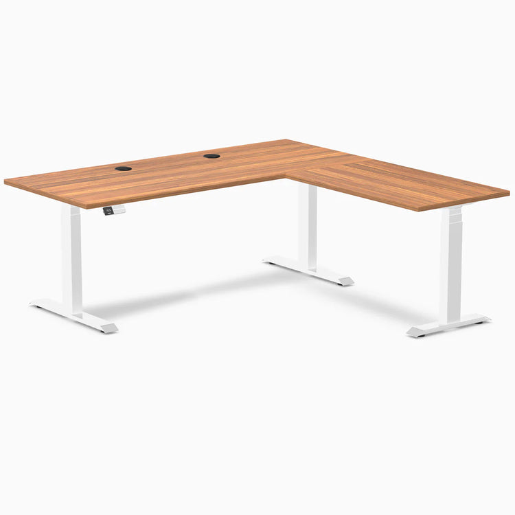 Prime Oak laminate L-shaped sit-stand desk with white adjustable legs and two black grommets.