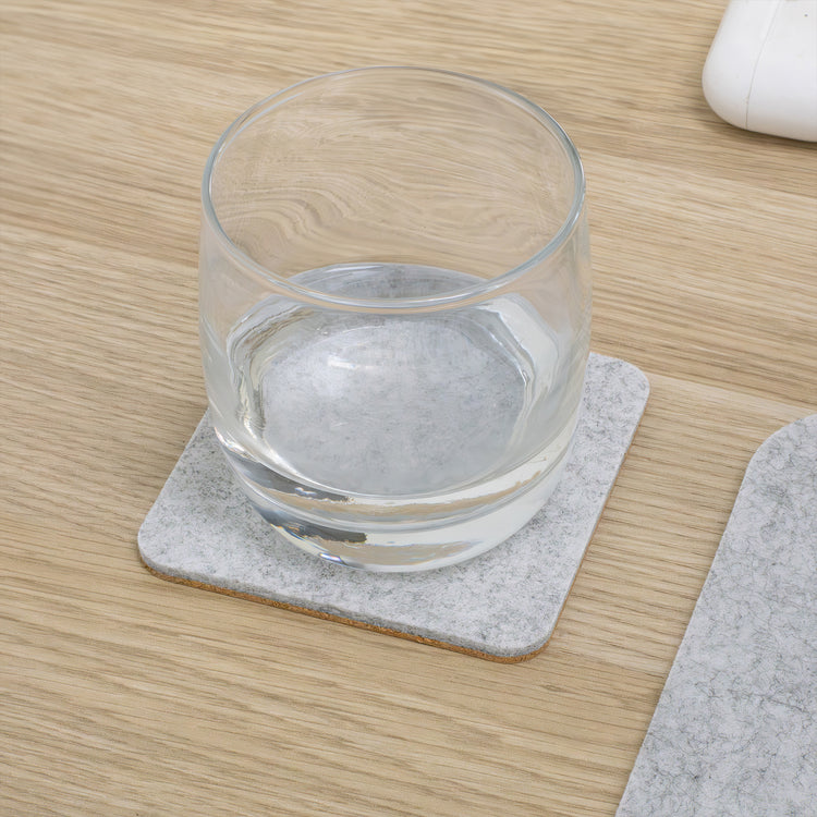 Square grey felt and cork coaster with a glass of water on a light wood desk.