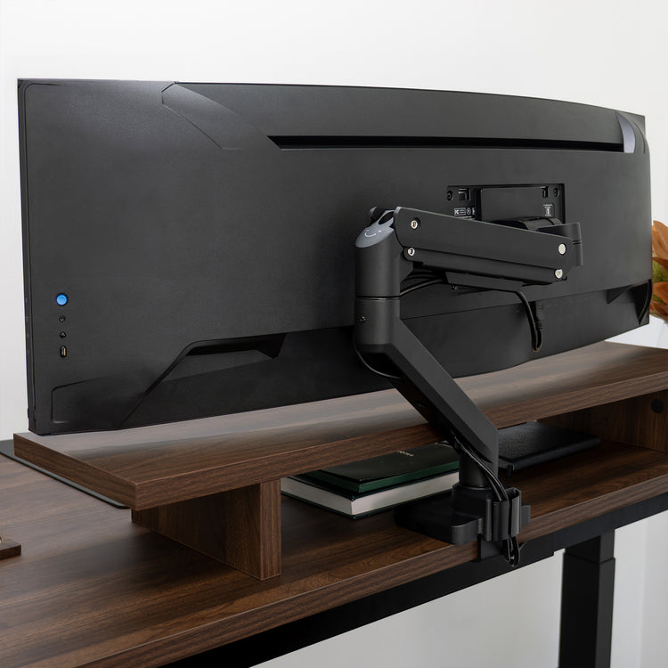 Black Desky Heavy Duty Ultrawide Monitor Arm securely holding a large ultrawide monitor on a dark wood desk.