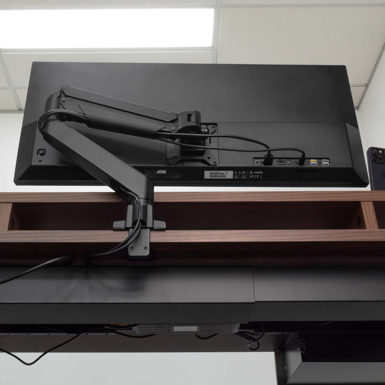 Black Desky Single Monitor Arm securely mounted to a dark wood desk, supporting a large black monitor.