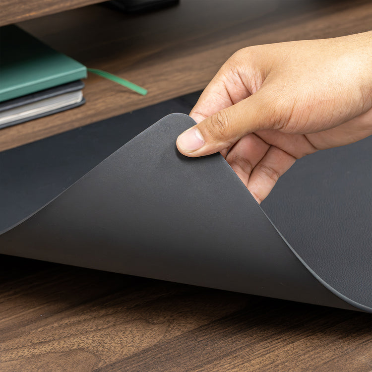 Hand lifting a corner of a large, dark gray Desky leather desk mat on a wooden desk.
