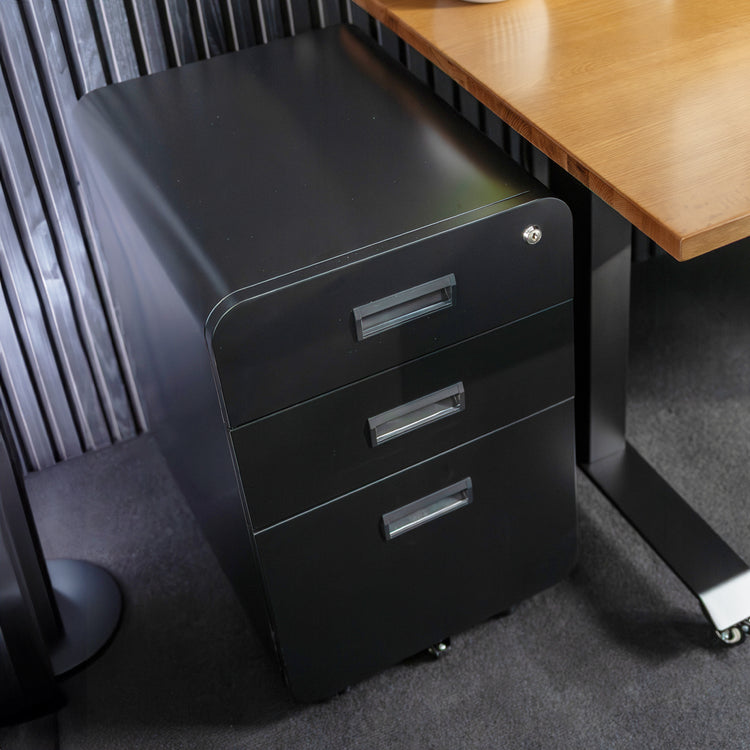 Desky 3 Drawer Mobile Filing Cabinet in black powder-coated steel with rounded edges, sitting under a wooden desk.