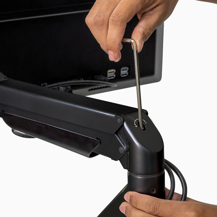 Hands adjust a black Desky Dual Monitor Arm with an Allen wrench, showing its sleek design and cable management.