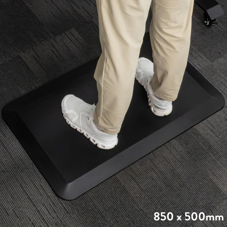 Black Desky anti-fatigue standing mat with a rugged surface and bevelled edge, shown with feet in white shoes.