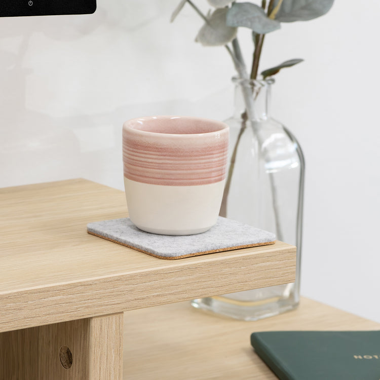 Light grey felt coaster with a pink and white ceramic mug on a light wood desk.
