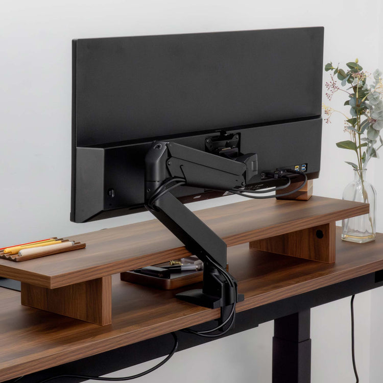 Black Desky Single Monitor Arm securely mounted to a dark wood desk, supporting a large black monitor.