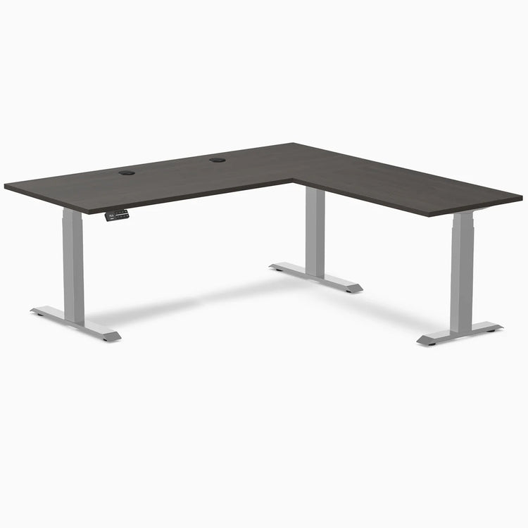 Burnished wood L-shape laminate sit-stand desk with gray frame and grommets.