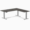 Burnished wood L-shape laminate sit-stand desk with gray frame and grommets.