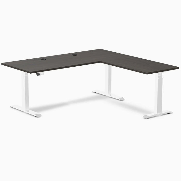 Burnished wood laminate L-shaped sit-stand desk with white adjustable legs and grommets.