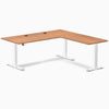 Prime Oak laminate L-shaped sit-stand desk with white adjustable legs and two black grommets.