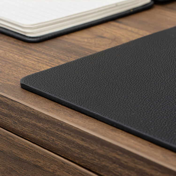 Desky black leather desk mat with pebbled texture on a wooden desk, notebook in background.