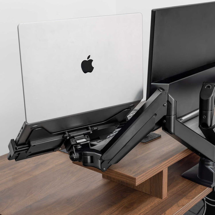 Black Desky Expandable Laptop Mount holding a silver MacBook, attached to a monitor arm on a wooden desk.