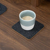 Square dark gray felt and natural cork coaster with a light blue and white mug on a wooden desk.