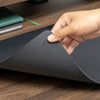 Hand lifting a corner of a large, dark gray Desky leather desk mat on a wooden desk.