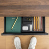 Black steel Desky under desk drawer with a green notebook, pens, and AirPods, seen from above.
