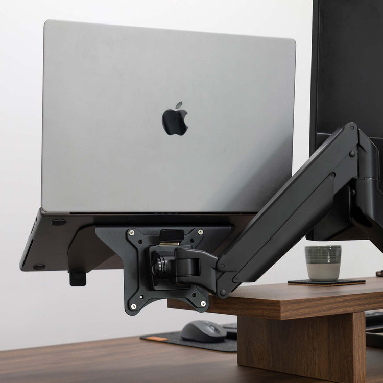 Desky Universal Laptop Mount in black, holding a silver MacBook, attached to a monitor arm on a wooden desk.