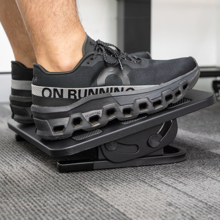 Person's feet in black running shoes resting on a black, tiltable Desky Classic Foot Rest with a non-skid surface.