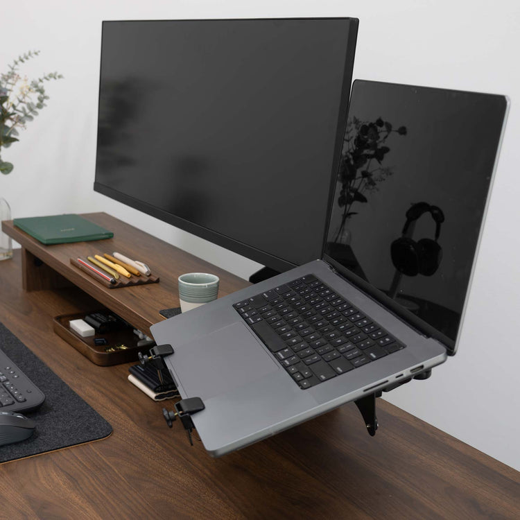 Desky Universal Laptop Mount in black, holding a silver laptop next to a monitor on a wooden desk.