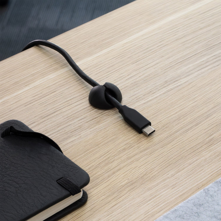Black Desky Cable Organizer clip holding a USB-C cable on a light wood desk next to a black notebook.