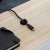 Black Desky Cable Organizer clip holding a USB-C cable on a light wood desk next to a black notebook.