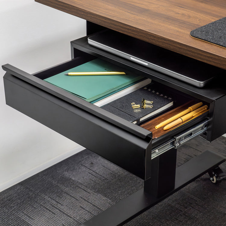Black steel Desky under-desk drawer, open and filled with notebooks, pens, and a laptop, attached to a dark wood desk.