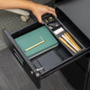 Hand opening the top drawer of a black steel Desky 3-drawer mobile filing cabinet, revealing an organizer tray with office supplies.