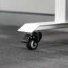 Black polyurethane locking caster wheel attached to a white steel desk leg on a gray carpet.