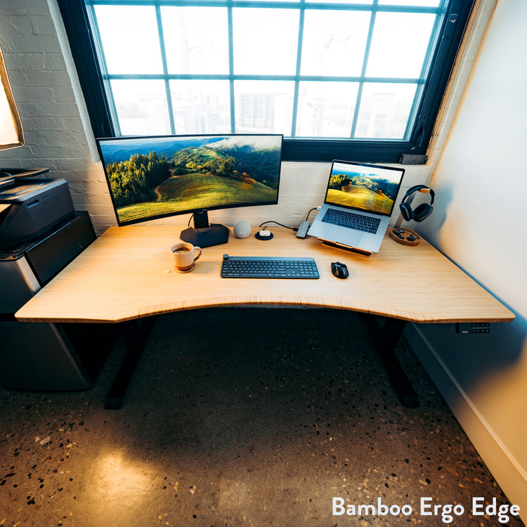 Light bamboo Desky Dual Ergo Edge sit-stand desk with a curved front, black frame, monitor, and laptop.