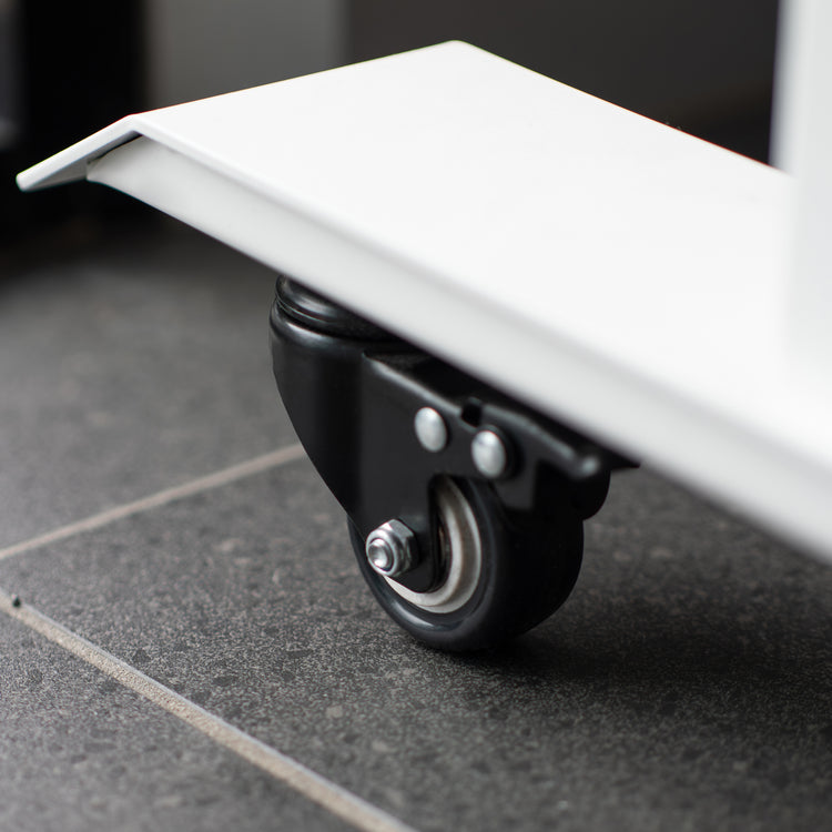 Close-up of a black polyurethane Desky lockable desk caster wheel attached to a white desk leg on a gray floor.