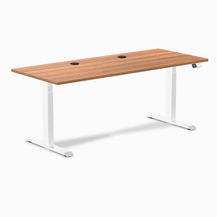 Desky Dual Prime Oak laminate sit-stand desk, 70.9x29.5 inches, with white frame and two black grommets.
